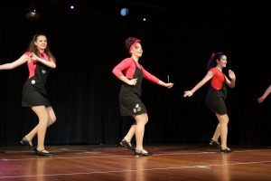 Three girls in black overalls tap dance at concert.