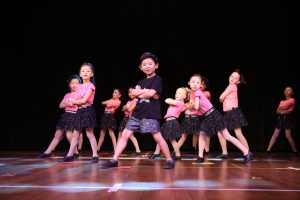 A young boy dressed in black dances in the foreground while in the backgroun a large number of girls in pink dance